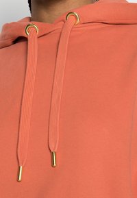 Miękki, koralowy hoodie o teksturowanej powierzchni, z złotymi oczkami i dopasowanymi sznurkami.