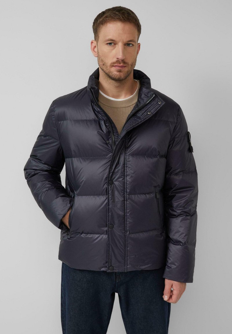 Mornarska puffer jakna s horizontalnim šavom, visokim ovratnikom, prednjim patentnim zatvaračem i kopčama na dugmad. Tkanina ima sjajnu, glatku teksturu.