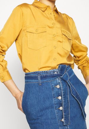 Kobieta ubrana w musztardowożółtą koszulę zapinaną na guziki z kieszeniami na patki oraz niebieską dżinsową spódnicę z wysokim stanem, zapinaną z przodu na guziki i związaną paskiem.
