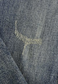 Denimstof in donkerblauw met een zichtbare vervaagde vlek en rafelige textuur, met een diagonale vouw die de binnenweving onthult.