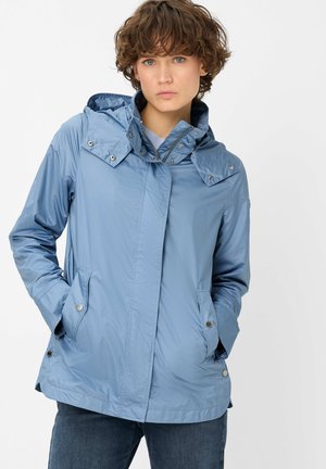 Frau trägt eine hellblaue Regenjacke mit Kapuze, Druckknopftaschen und Reißverschluss, Hände in den Taschen, steht vor einem schlichten weißen Hintergrund.