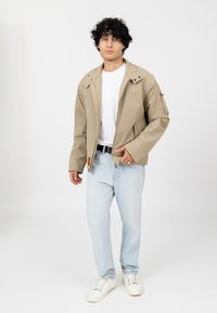 Jasnobeżowa kurtka z zapięciem na zamek, bocznymi kieszeniami i teksturowanym wykończeniem, w połączeniu z jasnoniebieskimi dżinsami i białymi trampkami.