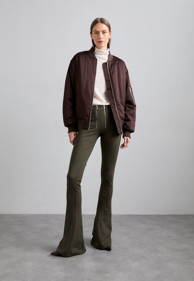 Brown bomber jacket with ribbed cuffs, worn over a cream turtleneck, paired with olive green flared pants in a smooth fabric.
