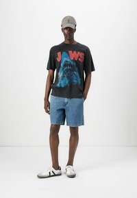 Grå grafisk T-skjorte med "JAWS" i røde bokstaver og haid design, sammen med blå denimshorts. Hvite sneakers med svarte detaljer.