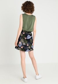 Top senza maniche verde oliva abbinato a una gonna nera con stampe tropicali colorate e vita arricciata, indossato con sneakers bianche.