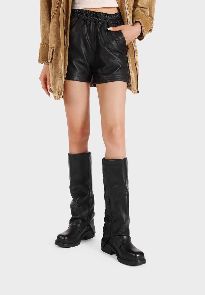 Pantaloni corti neri in pelle con una cintura elasticizzata abbinati a stivali neri in pelle alti fino al ginocchio con cerniere e un tacco spesso.