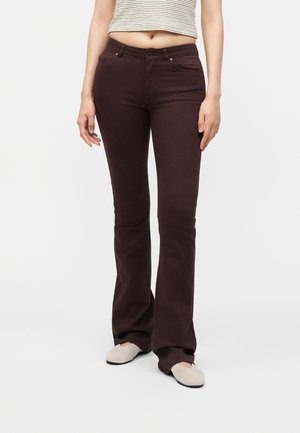 Kvinde iført mørkebrune bukser med svaj, beige flade sko og en ærmeløs crop top med vandrette striber, stående mod en ensfarvet baggrund.