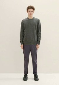 Νέος με σγουρά μαλλιά στέκεται μπροστά φοράει γκρίζο πουλόβερ, σκούρα παντελόνια με μανσέτες και μαύρα δερμάτινα παπούτσια με κορδόνια σε απλό φόντο.