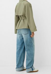 Olivově zelený kabát se stahováním v pase a peplum designem, v kombinaci se světle modrými oversized džíny s mírně vybledlou texturou a ohrnovanými lemy.