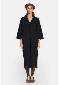 Czarna sukienka do kolan z dekoltem w kształcie litery V, z rękawami o długości trzech ćwierć. Modelka ma na sobie brązowe buty i pasującą opaskę na głowę.