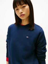 Jeune femme avec des cheveux noirs lisses portant un sweat-shirt bleu marine avec un petit logo rouge, blanc et bleu sur fond blanc.