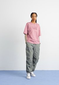 Roza bombažna majica s kratkimi rokavi z napisom "New Balance 1906" v temni barvi, v kombinaciji z ohlapnimi olivno zelenimi vojaškimi hlačami in belimi športnimi copati.