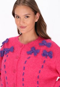 Cardigan roz cu țesătură texturată; dispune de fundițe din fetru mov și închidere contrastantă cu nasturi. Cusătura decorativă se desfășoară vertical pe față.