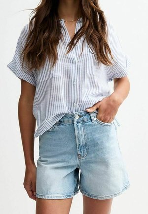 Kvinde iført en hvid, kortærmet skjorte med blå striber og lyseblå denimshorts, med den ene hånd i lommen.