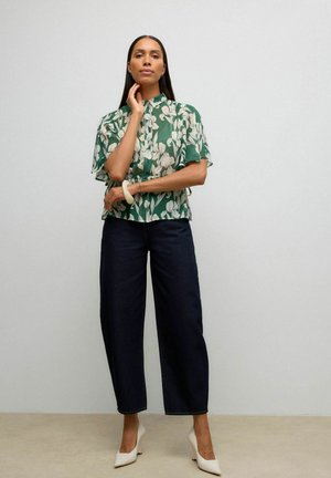 Femme portant un chemisier vert à fleurs à manches courtes, un pantalon large sombre, des talons blancs et un bracelet blanc, debout contre un mur uni.