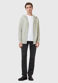Felpa grigia chiara con zip, realizzata in materiale morbido, indossata sopra una maglietta bianca, abbinata a jeans scuri e stivali neri.