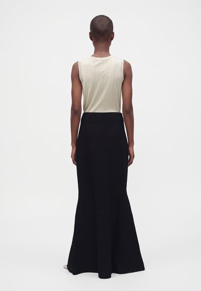 Person with short hair wearing a sleeveless beige top and long black skirt, standing facing away against a plain white background.