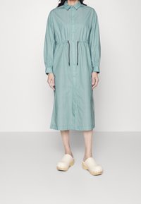 Donna che indossa un vestito midi azzurro chiaro con bottoni, vita con cordino e maniche lunghe, abbinato a zoccoli di legno beige.