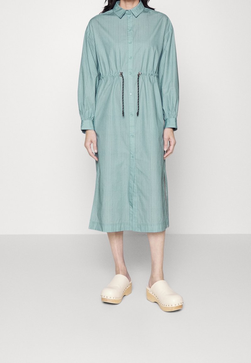 Donna che indossa un vestito midi azzurro chiaro con bottoni, vita con cordino e maniche lunghe, abbinato a zoccoli di legno beige.