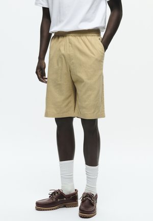 Person iført beige knælange shorts, hvide sokker, mørkebrune snørebåndssko og en hvid kortærmet skjorte med hænderne i lommerne.