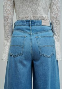 Vista trasera de una persona que lleva unos vaqueros azules de pierna ancha y cintura alta, y una blusa blanca ajustada de manga larga con encaje y patrón floral.