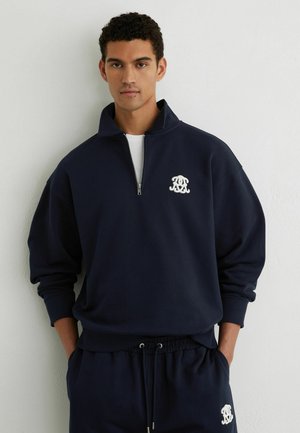 Jeune homme portant un sweat-shirt bleu marine à demi-fermeture éclair et un pantalon assorti avec des logos blancs brodés, debout contre un mur blanc.