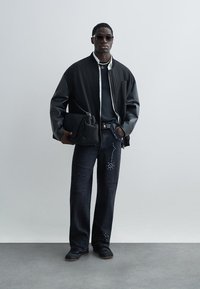 Άνδρας που φορά μαύρα γυαλιά ηλίου, μαύρο μπουφάν μπόμπερ με δερμάτινα μανίκια, σκούρα τζιν με σχέδια από στρας, κρατώντας μαύρη τσάντα, όρθιος.