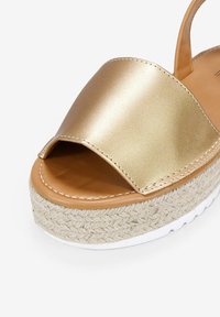 Sandalia abierta de dedo en metal dorado, que presenta una amplia tira, plataforma de yute natural trenzado y suela de goma blanca.