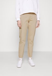 Beige bomullschinos med rak passform, parat med vita sneakers och en ljusrosa huvtröja, uppvisad mot en enfärgad bakgrund.