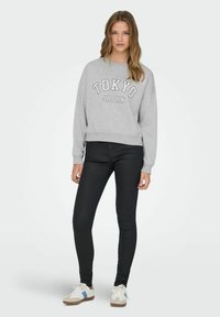 Ung kvinde iført en grå "TOKYO JAPAN" sweatshirt, sorte skinny jeans og hvide sneakers med blå striber, stående mod en almindelig baggrund.