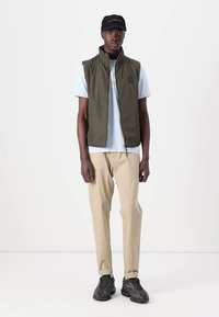 Olive green sleeveless vest, light blue t-shirt, cream pants, black sneakers, and cap, all in a casual fit against a plain background.