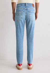 Persoon in lichtblauwe denim jeans en beige sneakers met rode accenten, staande tegen een effen witte muur.