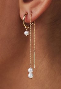 Boucles d'oreilles en or avec une chaîne ornée de deux perles blanches et d'un accent en métal droit. Le design met en avant une simplicité élégante et un contraste.