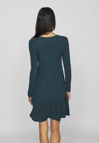 Rippkleid mit langen Ärmeln in dunklem Teal und gestuften Saum. Weicher Strickstoff, taillierter Oberkörper und ausgestellt Rock.