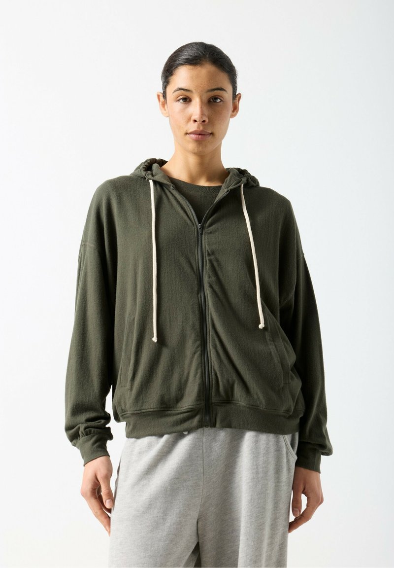 Sudadera con capucha con cremallera de color verde oliva y tela texturizada, con capucha ajustable mediante cordón y bolsillos delanteros, combinada con pantalones deportivos grises.