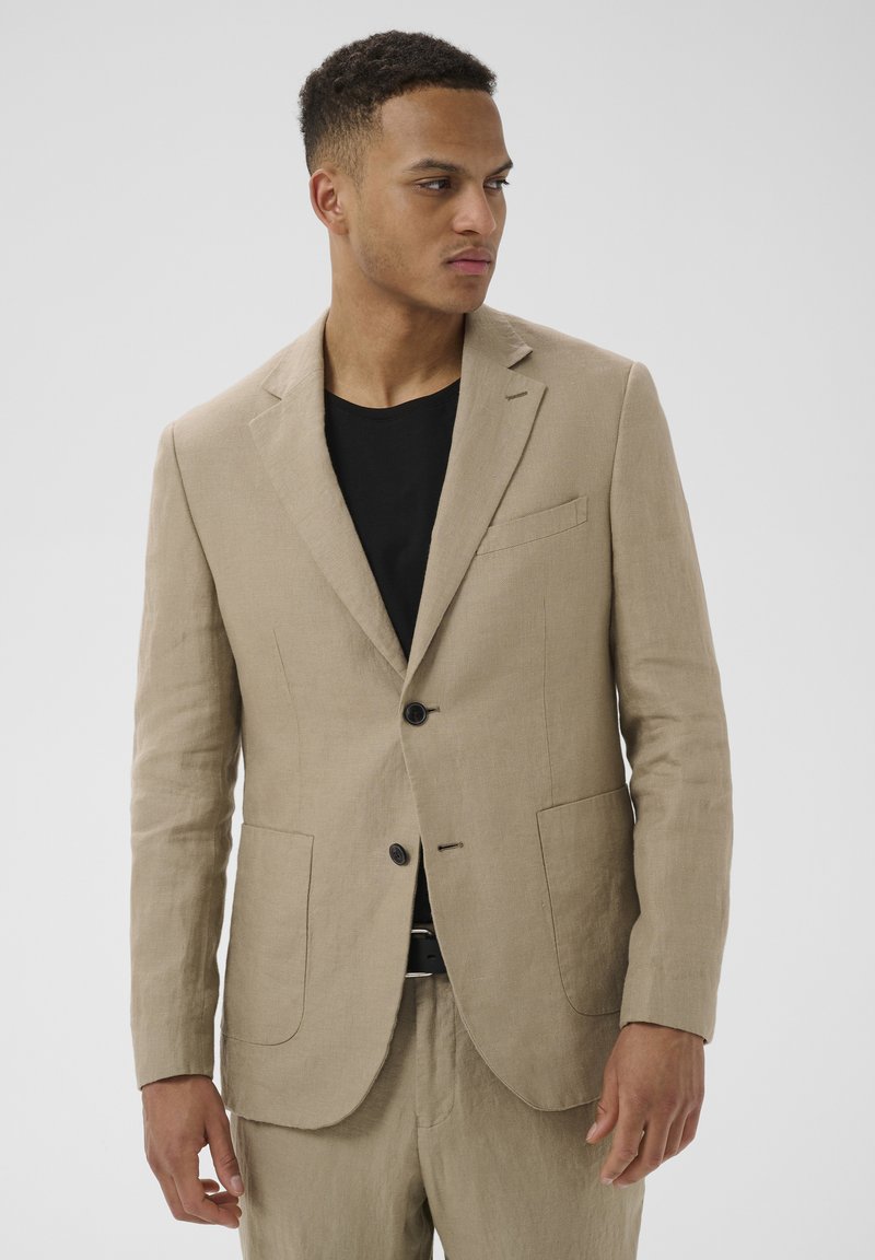 Homme portant une veste de costume en lin beige et un pantalon assorti sur une chemise noire, regardant vers sa gauche devant un fond uni.