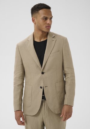 Mann trägt eine beige Leinenanzugsjacke und passende Hose über einem schwarzen Hemd, schaut nach links vor einem schlichten Hintergrund.