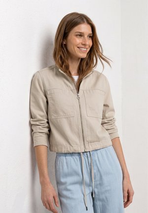 Ung kvinde smiler og læner sig op ad en hvid væg iført en beige jakke med lynlås og lyseblå bukser med snøre.
