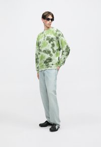 Πράσινο φούτερ tie-dye με γύρο λαιμό και μακριά μανίκια, συνδυασμένο με ανοιχτό μπλε τζιν και μαύρα παπούτσια, σε απλό φόντο.