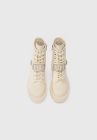 Bottines en cuir synthétique couleur crème avec un laçage à l'avant, une sangle en métal argenté marquée "GUESS", et un design de semelle chunky.