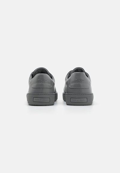 Szürke bőr sneaker, lekerekített szélekkel, texturált gumitalppal, dombornyomott logóval a sarkán, és minimalista dizájnnal.