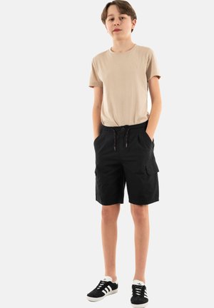T-shirt en coton beige, shorts cargo noirs avec poches latérales, taille élastique et cordon rouge. Baskets noires avec des rayures blanches.