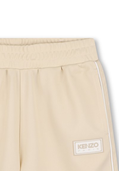 Shorts de sport beiges en tissu texturé. Taille élastique avec cordon de serrage, poches latérales et un patch logo blanc à l'avant.