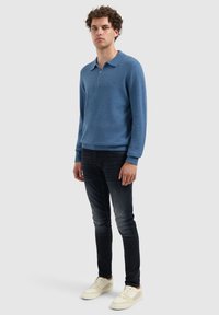 Blauwe gebreide pullover met een zachte textuur, met ritssluiting bij de kraag en lange mouwen, gecombineerd met donkere jeans en lichtgekleurde sneakers.