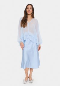 Blusa trasparente azzurro chiaro con volant strati, abbinata a una gonna midi in satin dello stesso colore. I mule con fiocco argentato completano il look.