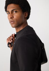 Schwarzes Langarmshirt mit glatter Textur und Schulternähten. Das Model hält einen schwarzen Riemen einer kleinen Tasche über der Schulter.