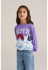 Lilla sviiter, millel on valges kirjas "ALPES", sinise ja punase mägise stseeniga. Kombineeritud helesiniste teksadega, mis on kaunistatud kivega.