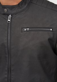 Chaqueta de cuero negro con cuello alto, cierre frontal de cremallera y un bolsillo lateral con cremallera y herrajes en plata. Textura suave y diseño contorneado.