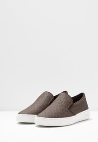Chaussures à enfiler marron avec dessus en tissu texturé et semelle en caoutchouc blanc. Présente un motif subtil et un design à bout rond. Aucun matériel visible.