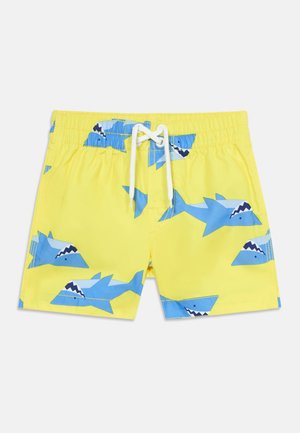 Short de bain jaune avec imprimés de requins dessin animé bleus et taille élastiquée avec cordon blanc sur fond blanc.
